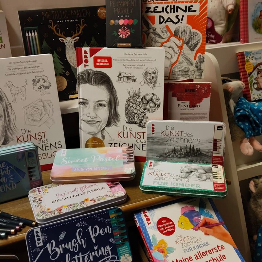 Eine Auswahl an Zeichenbüchern ist auf einem Regal ausgestellt. Titel umfassen 'Die Kunst des Zeichnens' und 'Zeichnen Das!'. Ein Teddybär befindet sich rechts von den Büchern. Eine Schachtel Buntstifte steht vor den Büchern auf dem Tisch.