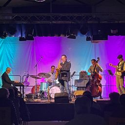 Eine Band tritt auf der Bühne mit lila und blauen Lichtern auf. Der Mann, der die Saxophon spielt, steht in der Mitte. Rechts spielt ein Mann das Cello und links spielt ein Mann das Schlagzeug.