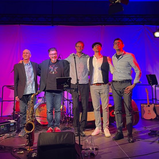 Fünf Musiker stehen auf einer Bühne mit Musikinstrumenten hinter ihnen. Einer hält eine Saxophon und sie tragen alle Freizeitkleidung. Sie sind von Bühnenlichtern beleuchtet.