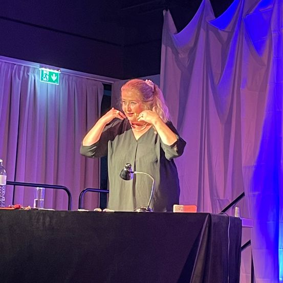 Eine Frau steht an einem Schreibtisch und spricht in ein Mikrofon. Sie trägt ein graues Oberteil und hat einen Schal um den Hals. Hinter ihr steht ein Tisch mit einer Lampe, einer Wasserflasche und einem Glas. Vorhang und eine lila Beleuchtung sind im Hintergrund.