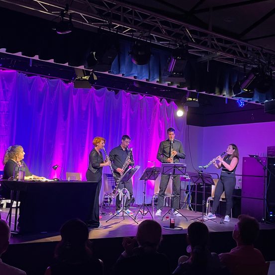 Eine Gruppe von Musikern spielt auf der Bühne mit lila Beleuchtung. Sie spielen Instrumente, darunter Klarinette, Oboe und Saxophon. Ein Publikum beobachtet sie.