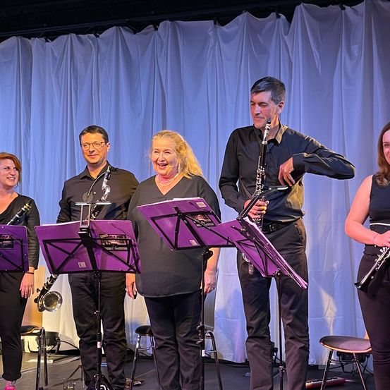 Fünf Musiker stehen lächelnd auf der Bühne und halten Musikinstrumente in den Händen. Sie sind alle in Schwarz gekleidet. Hinter ihnen befindet sich ein weißer Vorhang.