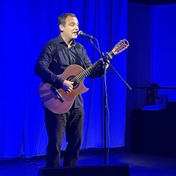 Ein Mann steht auf der Bühne, hält eine akustische Gitarre und singt in ein Mikrofon. Blaue Vorhänge sind hinter ihm.