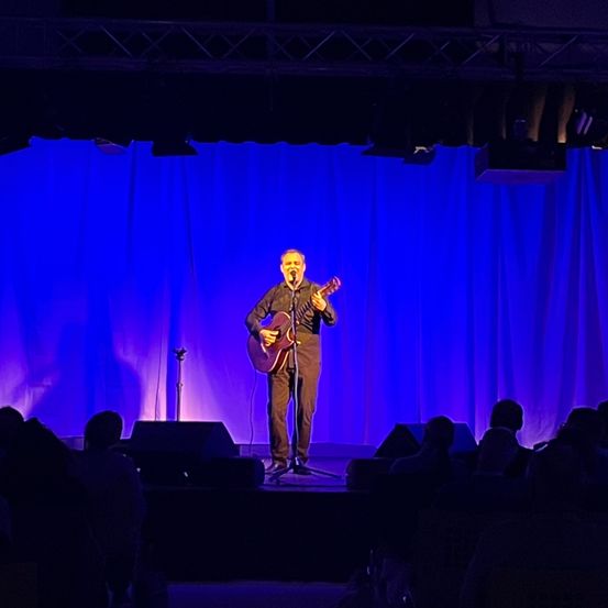 Ein Mann steht mit einer Gitarre auf der Bühne und tritt für ein Publikum unter blauem Licht auf.