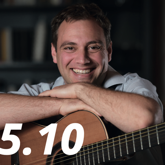 Ein lächelnder Mann mit Brille hält eine Gitarre mit verschränkten Armen. Auf der Gitarre steht die Nummer 5.10.