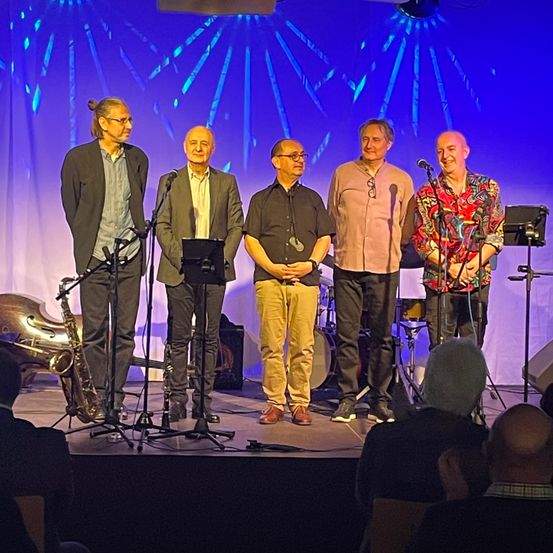 Fünf Männer stehen auf einer Bühne mit Musikinstrumenten vor ihnen. Ein Saxophon ist links. Hinter ihnen sind ein Schlagzeug und Lautsprecher aufgebaut. Die Bühne hat einen blauen Hintergrund mit einem Lichtdesign. Das Publikum sitzt vor der Bühne.