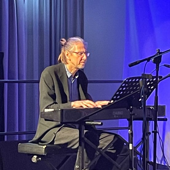 Ein älterer Mann mit Brille sitzt auf der Bühne an einem Klavier, mit einem Mikrofonstativ vor ihm.