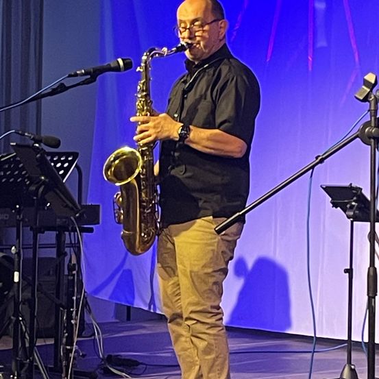Ein Mann spielt auf der Bühne ein Saxophon mit einem Mikrofon vor sich, trägt eine Brille und eine Uhr. Hinter ihm stehen ein Klavier und andere Ausrüstung auf der Bühne.