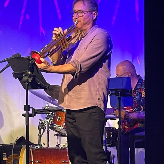 Ein Mann spielt Trompete auf der Bühne, mit einem Schlagzeuger hinter ihm. Die Bühne ist mit lila Lichtern beleuchtet.