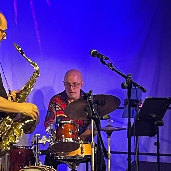 Zwei Musiker sind auf der Bühne mit einem blauen Hintergrund. Die Person auf der linken Seite spielt Saxophon, während die auf der rechten Seite Schlagzeug spielt.