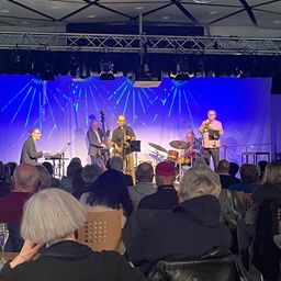 Eine Jazzband tritt auf der Bühne unter blauem Licht auf, mit Instrumenten wie Trompeten, Saxophonen und Schlagzeug. Das Publikum, bestehend aus älteren Erwachsenen, beobachtet aufmerksam von vorne.