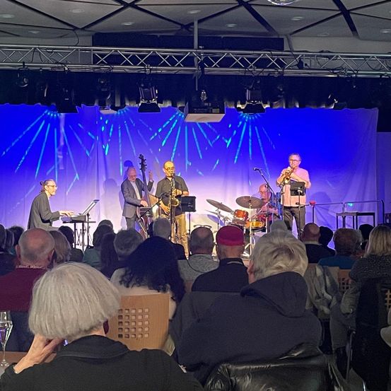 Eine Jazzband tritt auf der Bühne unter blauem Licht auf, mit Instrumenten wie Trompeten, Saxophonen und Schlagzeug. Das Publikum, bestehend aus älteren Erwachsenen, beobachtet aufmerksam von vorne.