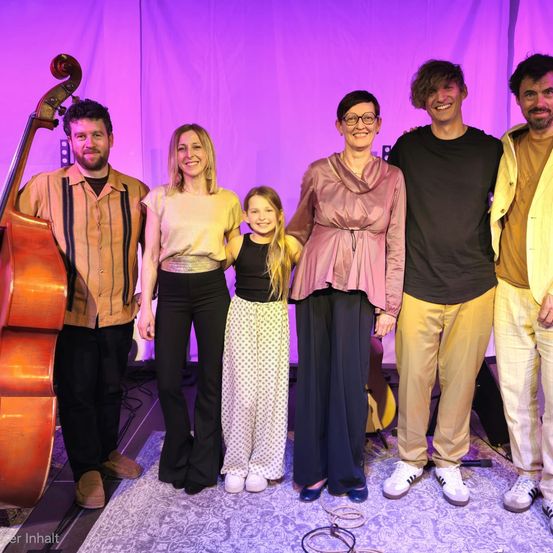 Eine Gruppe von Musikern und einem Kind posiert vor einem lila Hintergrund für ein Foto. Ein Mann spielt Cello, und andere tragen Brillen, Turnschuhe und Armbänder.