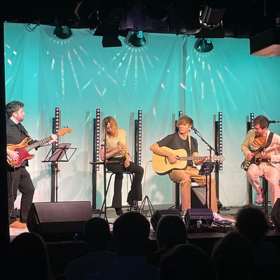 Vier Musiker spielen auf einer Bühne mit einem türkisfarbenen Hintergrund, wobei jeder ein Instrument spielt. Der Mann links spielt eine Bassgitarre, während die Frau in der Mitte ein Mikrofon hält. Zwei Männer sitzen auf beiden Seiten und spielen jeweils eine Gitarre und singen.