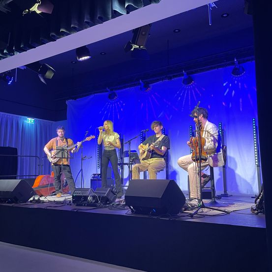 Eine Band spielt auf der Bühne mit Gitarren und Mikrofon unter blauem Licht. Das Publikum ist nicht sichtbar.