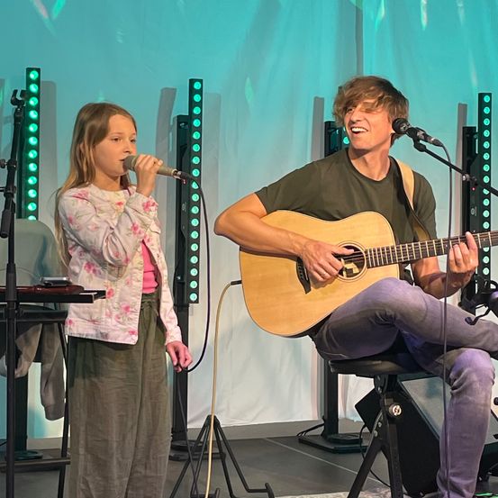 Ein junges Mädchen und ein Mann treten auf der Bühne auf. Das Mädchen hält ein Mikrofon, während der Mann die Gitarre spielt. Beide lächeln.