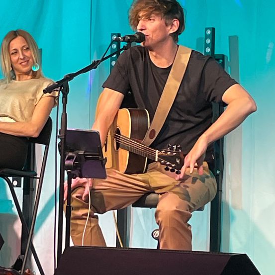 Ein Mann spielt Gitarre und singt in ein Mikrofon auf der Bühne, während eine Frau neben ihm lächelnd sitzt.