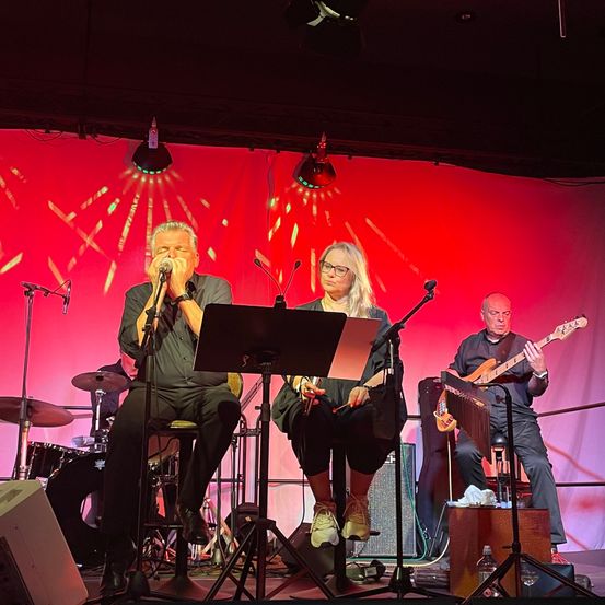 Eine Band tritt auf der Bühne mit rotem Hintergrund auf. Ein Mann singt, eine Frau spielt Flöte und ein Mann spielt Gitarre. Andere Instrumente und Lautsprecher sind ebenfalls vorhanden.