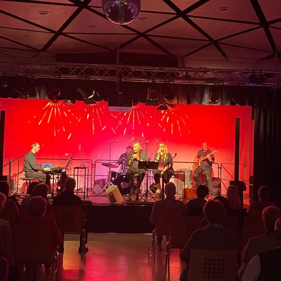 Eine Band spielt auf der Bühne mit rotem Hintergrund und Scheinwerfern, während das Publikum auf Stühlen sitzt.