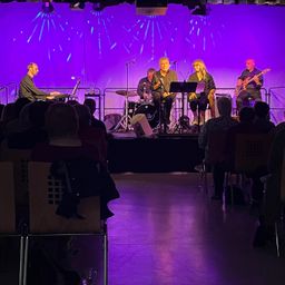 Eine kleine Band spielt auf einer Bühne mit lila Beleuchtung vor einem Publikum. Ein Mitglied spielt Klavier, während andere Gitarre und Schlagzeug spielen.