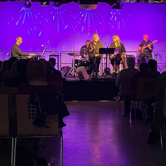Eine kleine Band spielt auf einer Bühne mit lila Beleuchtung vor einem Publikum. Ein Mitglied spielt Klavier, während andere Gitarre und Schlagzeug spielen.
