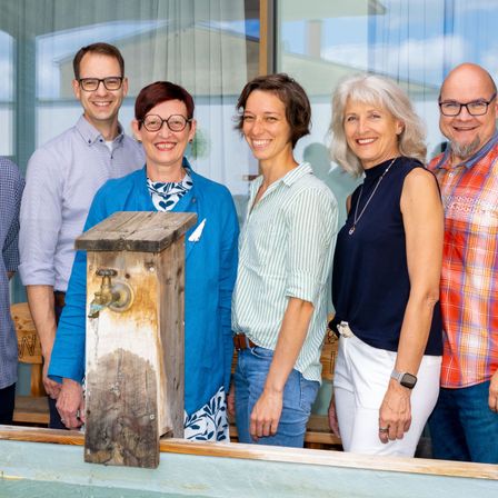 Fünf Personen stehen eng beieinander und lächeln für ein Foto, wobei ein hölzerne Nistkasten im Vordergrund steht. Die beiden Frauen tragen Blau und Weiß, während die Männer lila, karierte und karierte Hemden tragen.