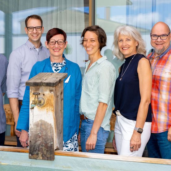 Fünf Personen stehen eng beieinander und lächeln für ein Foto, wobei ein hölzerne Nistkasten im Vordergrund steht. Die beiden Frauen tragen Blau und Weiß, während die Männer lila, karierte und karierte Hemden tragen.