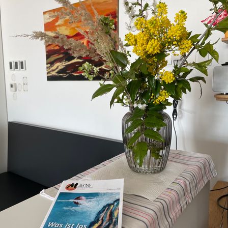Ein Tisch trägt eine klare Glasvase mit gelben Blumen. Ein Magazin mit dem Titel 'Was ist los' liegt auf dem Tisch. Hinter dem Tisch zeigt eine Wand ein Gemälde mit rotem und orangem Hintergrund.