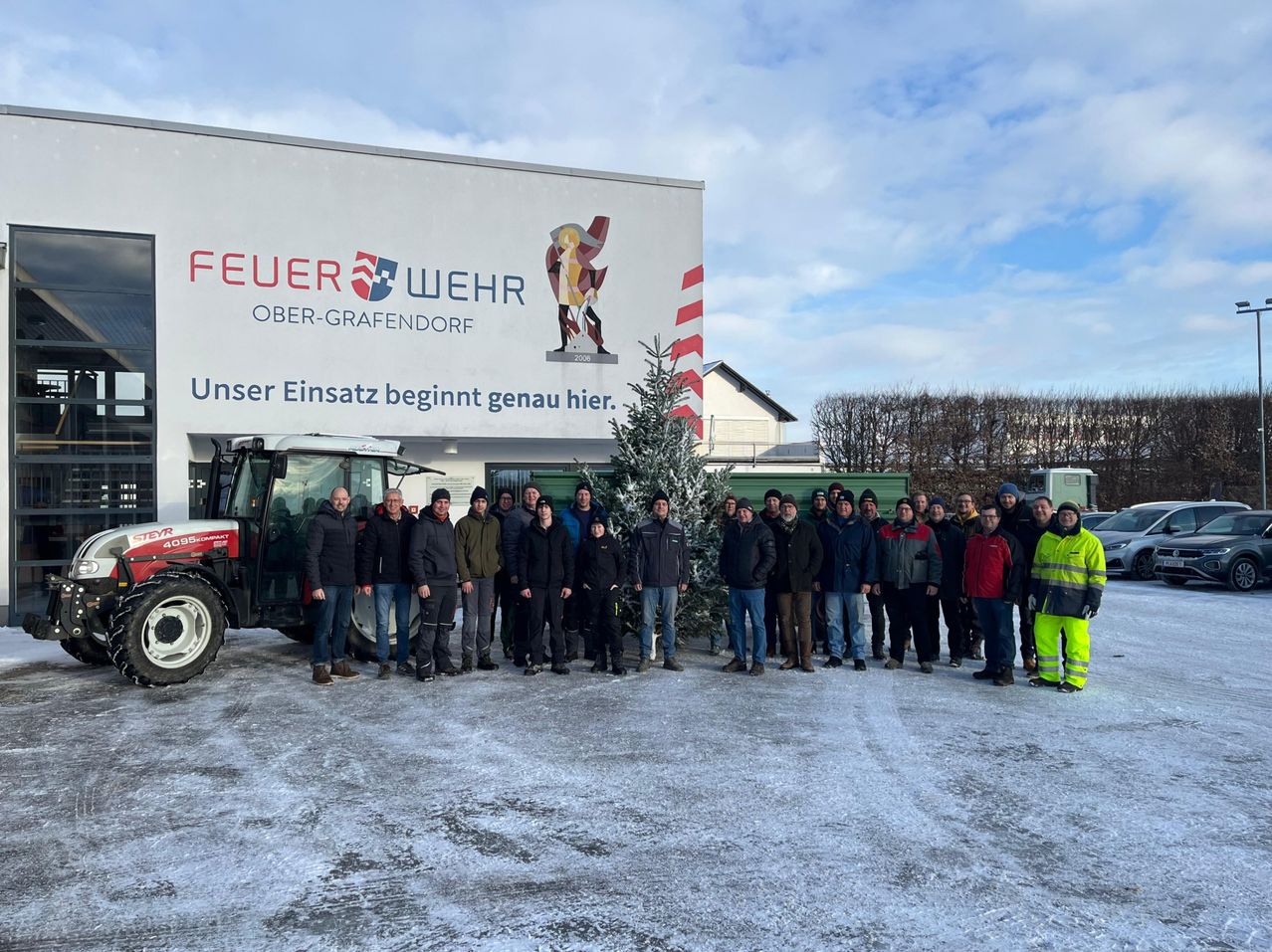 Eine Gruppe von Menschen steht vor einem Gebäude mit einem Traktor und einem Weihnachtsbaum. Das Gebäude trägt den Schriftzug 'FEUER WEHR OBER-GRAFENDORF'. Der Himmel ist bewölkt.