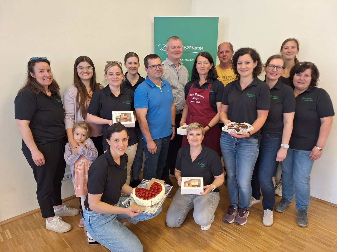 Eine Gruppe von Menschen posiert für ein Foto mit Kuchen und Gebäck. Einige tragen schwarze Uniformen, während andere Freizeitkleidung tragen. Ein grüner Banner ist im Hintergrund.