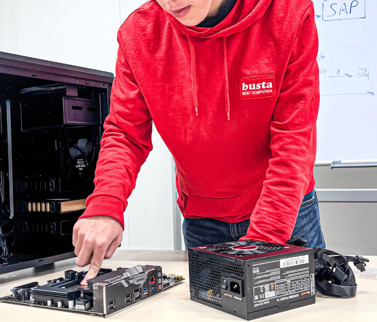 Eine Person in einer roten Kapuzenjacke arbeitet an einem Computer-Motherboard und Netzteil auf einem Schreibtisch.