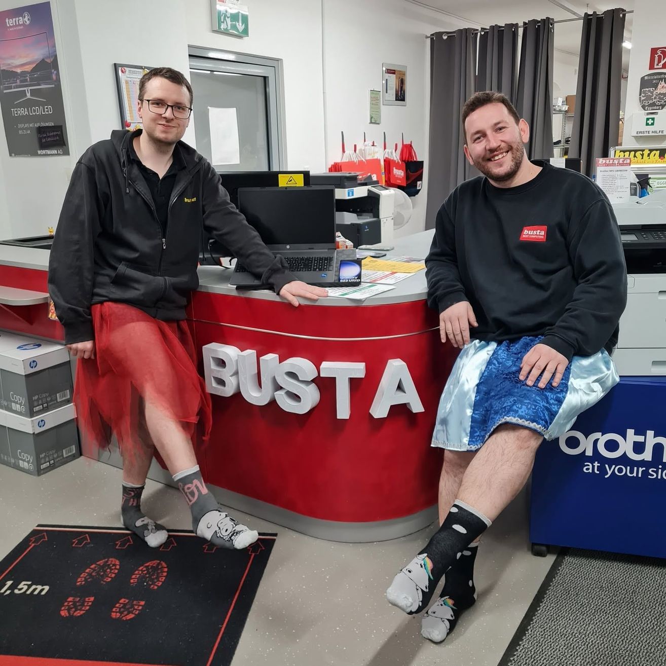 Zwei Männer in bunten Kostümen stehen hinter einer Theke mit dem Wort 'BUSTA' darauf. Ein Mann trägt einen roten Rock und Socken mit Cartoon-Figuren, während der andere blaue Shorts und Socken mit einem weißen Design trägt. Sie lächeln und posieren für ein Foto.