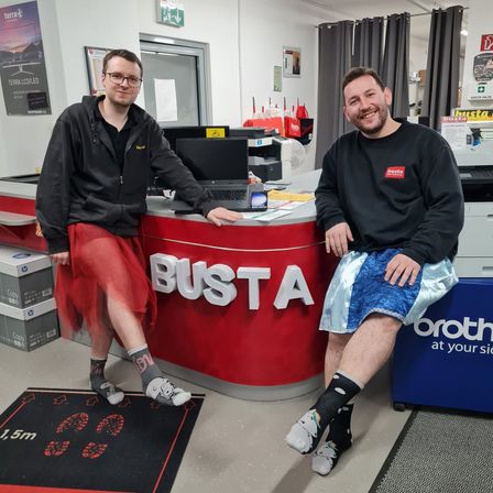 Zwei Männer in bunten Kostümen stehen hinter einer Theke mit dem Wort 'BUSTA' darauf. Ein Mann trägt einen roten Rock und Socken mit Cartoon-Figuren, während der andere blaue Shorts und Socken mit einem weißen Design trägt. Sie lächeln und posieren für ein Foto.