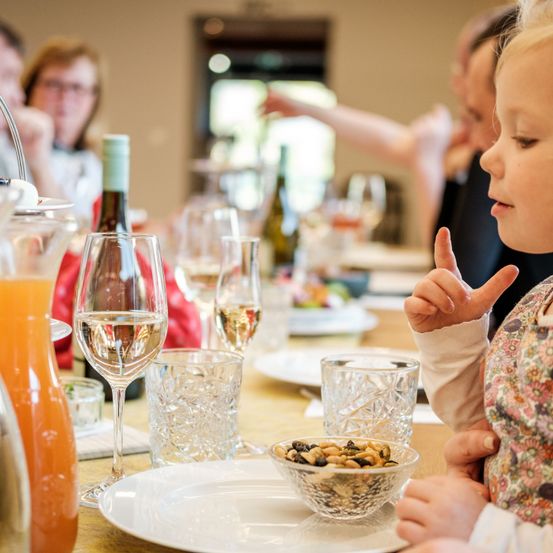 Ein Tisch für eine Mahlzeit mit Weinläsern, einer Schüssel mit Nüssen und einem kleinen Mädchen mit einem Zeichen. Andere Menschen sitzen hinter ihr.