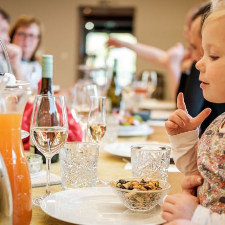 Eine Familie sitzt am Tisch, ein Kind hält einen Finger hoch, Wein- und Wassergläser, Flaschen und eine Schüssel mit Nüssen sind zu sehen.