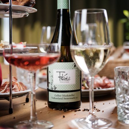 Ein Tisch mit einer Flasche Weißwein und zwei Weingläsern, eines voll und eines leer, neben einem Teller mit Fleisch und verschiedenen Gerichten.