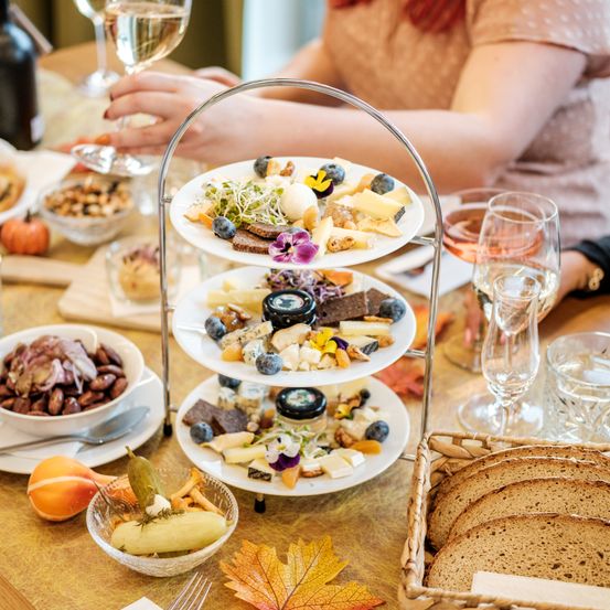 Ein Tisch mit einem dreistufigen Teller mit Käse, Brot und Obst sowie Weingläsern. Eine Frau sitzt am Tisch.