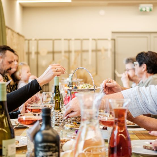 Eine Familie sitzt um einen Esstisch herum und genießt Essen und Getränke. Eine Frau greift nach einem Gericht auf einem gestapelten Ständer, während andere mit den Händen essen. Weingläser und Flaschen stehen auf dem Tisch, und sie scheinen sich in einer entspannten, geschlossenen Umgebung zu befinden.