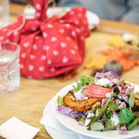 Eine Nahaufnahme eines Salats mit Apfel, Kürbis und Rotkohl auf einem Holztisch mit Herbstblättern und einer roten Geschenktüte.