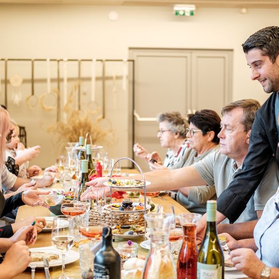 Eine Gruppe Erwachsener sitzt um einen Tisch herum, der mit Wein, Gläsern und Essen gefüllt ist. Ein Mann im Anzug lächelt und spricht mit der Gruppe.