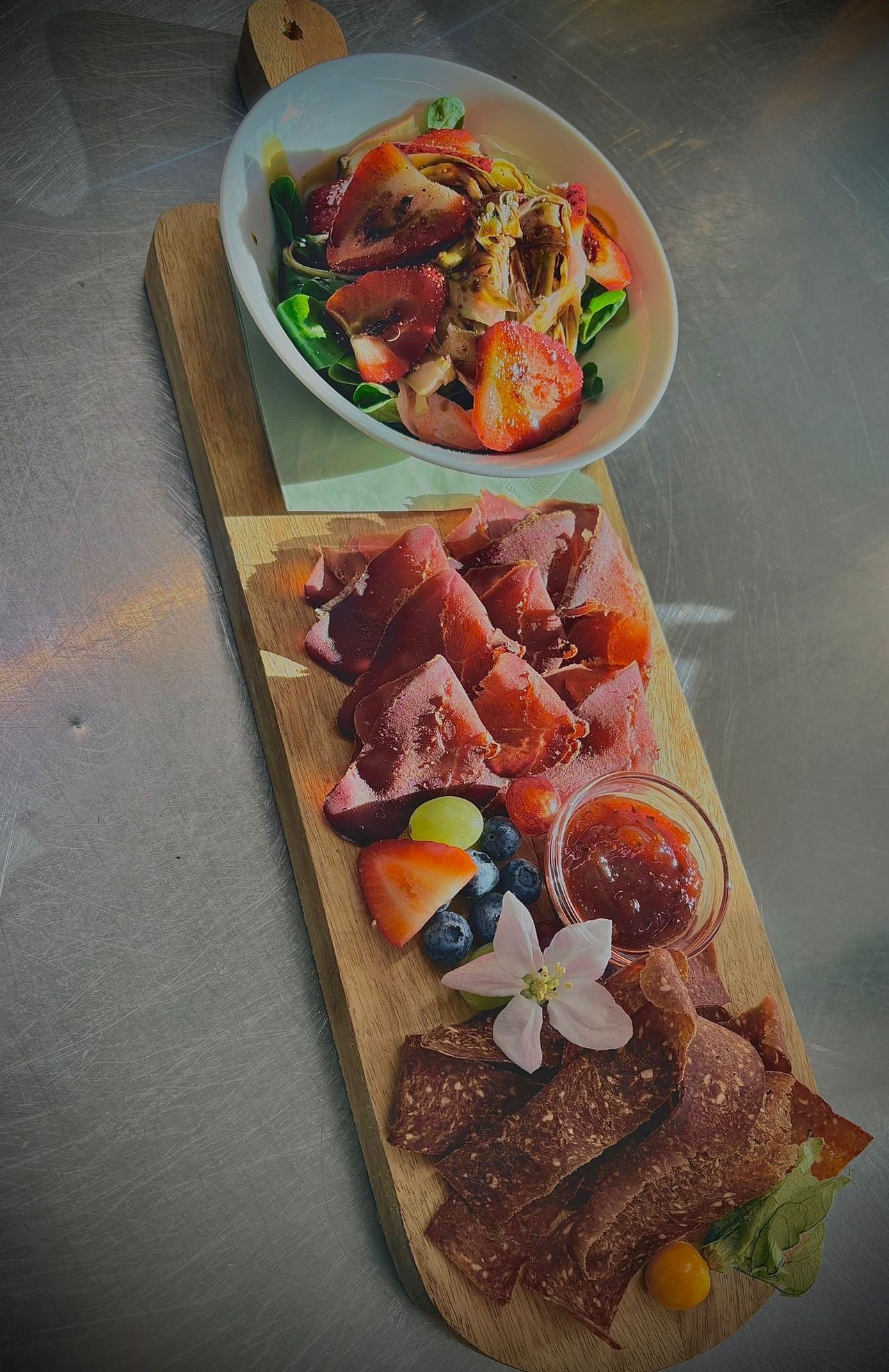 A wooden platter with assorted meats, a small bowl of salad, and fruits. A jar of jam and a white flower are also on the platter.