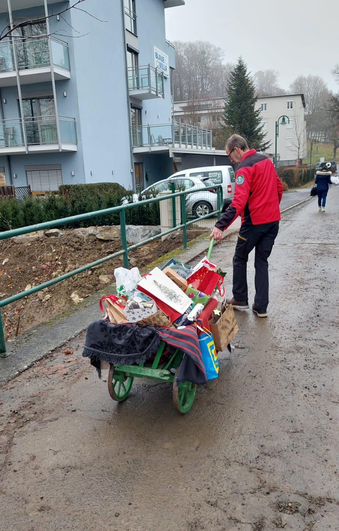 Ein Mann schiebt einen grünen Schubkarren voller verschiedener Geschenke und Gegenstände und geht auf einer nassen Straße. Hinter ihm ist eine grüne Brüstung und in der Ferne geht eine Person und Gebäude mit Balkonen.