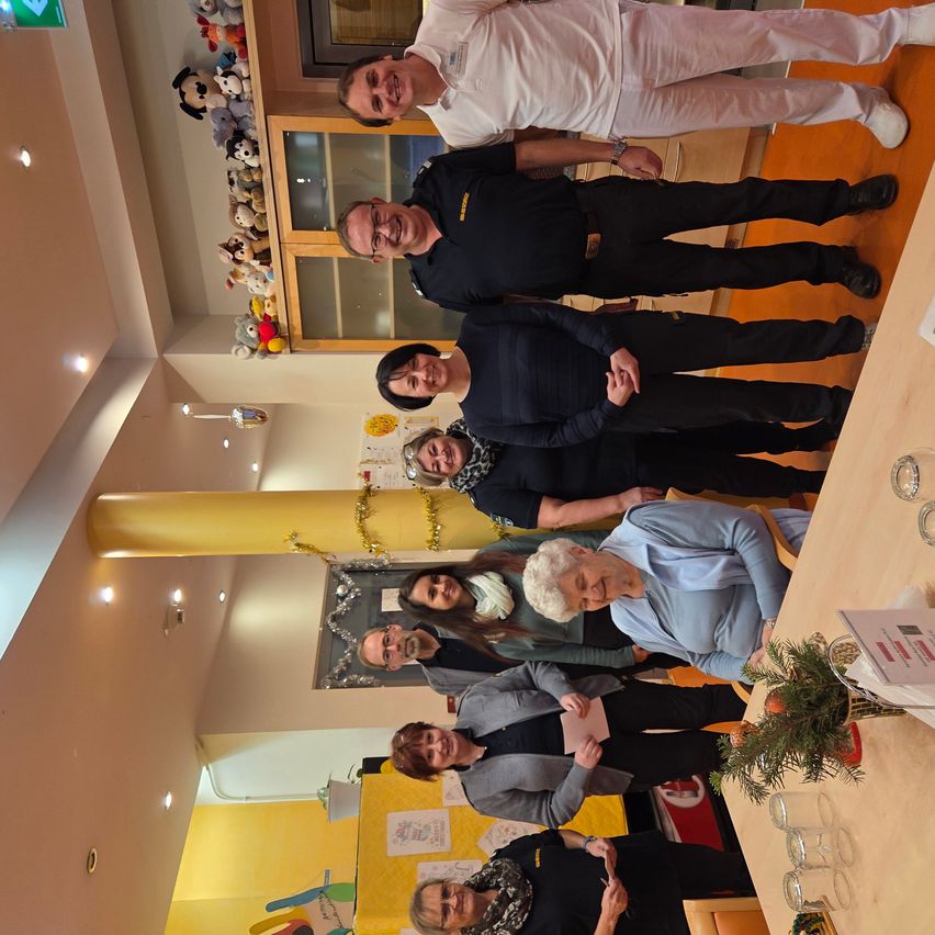 Eine Gruppe von Menschen steht zusammen in einem Raum, einige lächeln und andere posieren. Sie sind alle in Schwarz gekleidet, wobei eine ältere Frau ein hellblaues Shirt trägt. Es gibt einen Tisch mit einer Pflanze und Gläsern darauf.