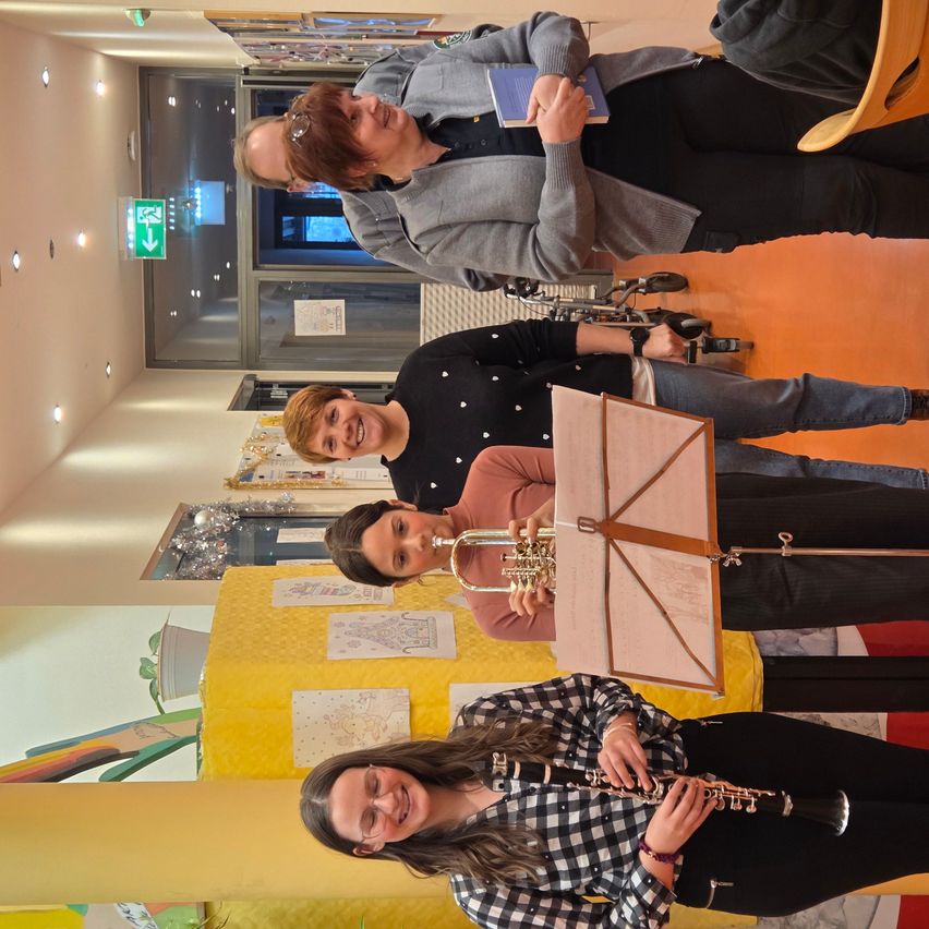 Fünf Personen stehen in einem Flur mit Musikinstrumenten. Zwei halten Instrumente, eine liest ein Buch und zwei lächeln.