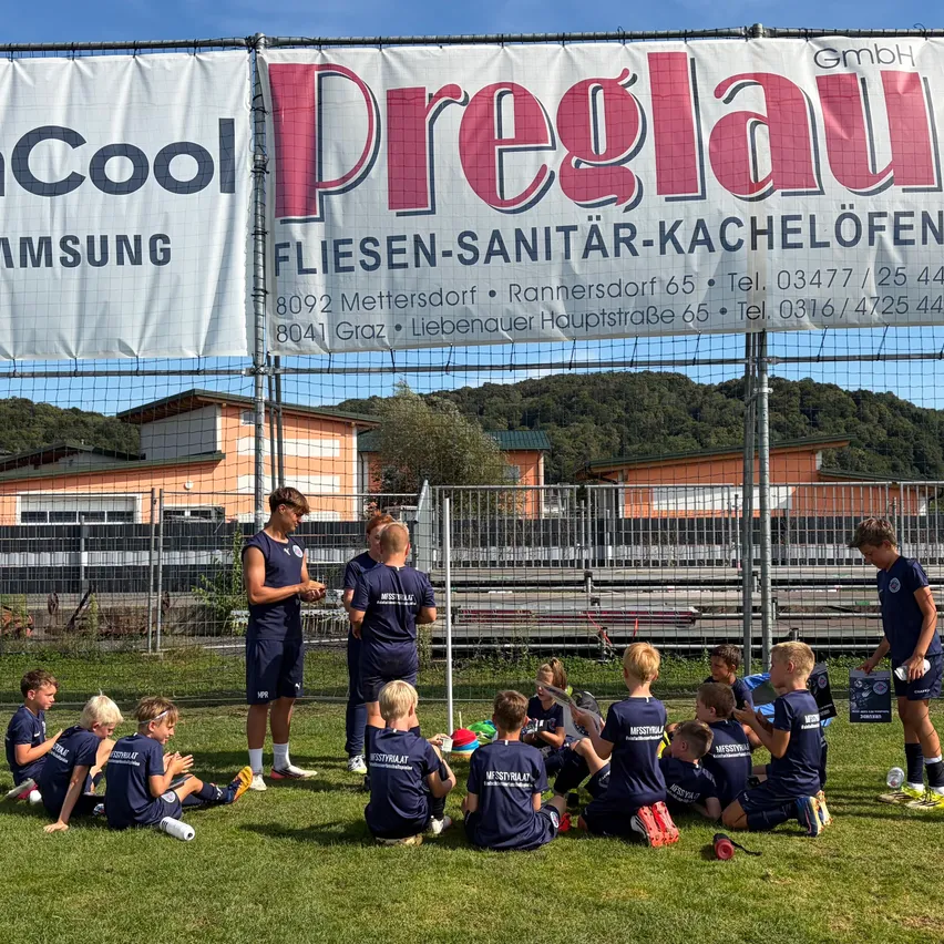 Eine Gruppe junger Fußballspieler in blauen Trikots versammelt sich auf einem Rasenfeld, vor ihnen steht ein Trainer. Dahinter befindet sich ein Banner mit dem Text 'Preglauer Fliesen-Sanitär-Kachelöfen'. Das Feld ist von einem Zaun umgeben, und im Hintergrund befinden sich Gebäude und Bäume.
