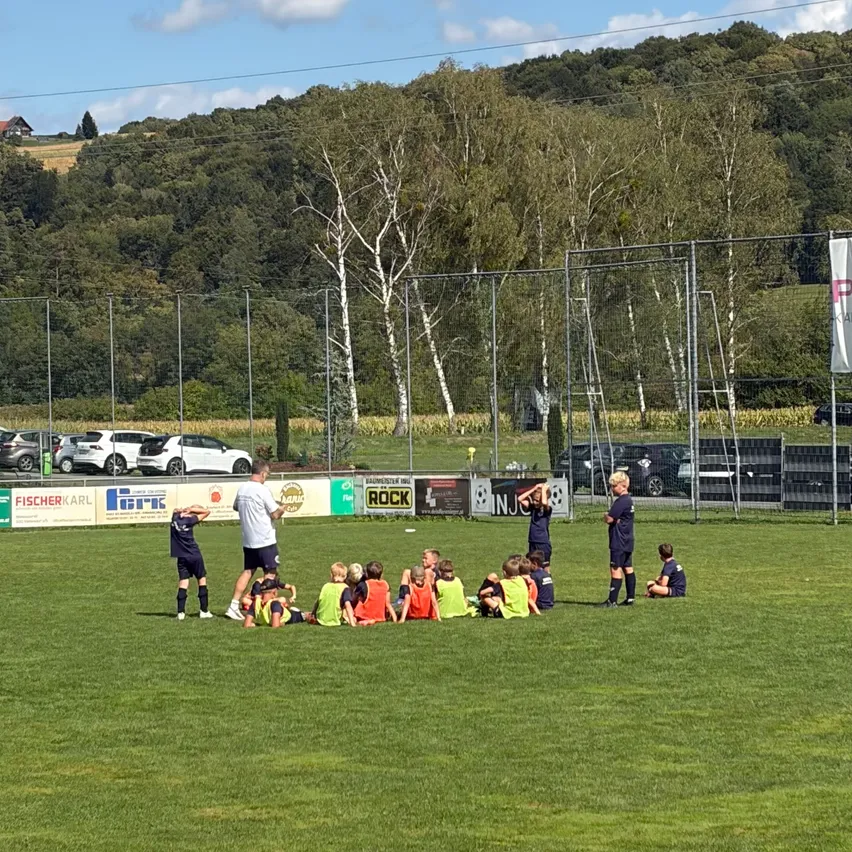 Eine Gruppe junger Fußballspieler versammelt sich auf einem Feld, während ein Trainer Anweisungen gibt. Das Feld ist von einem Zaun mit Bannern umgeben. Im Hintergrund befinden sich Bäume und geparkte Autos.