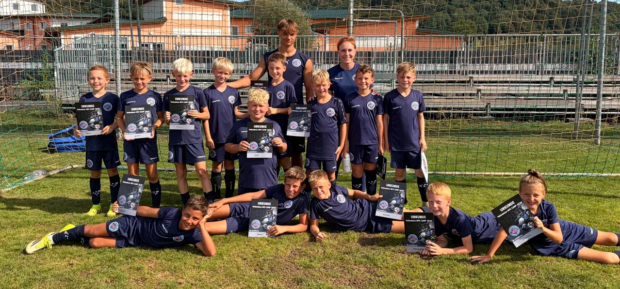 Eine Gruppe junger Fußballspieler, einige stehend und andere auf dem Rasen liegend, halten Urkunden in den Händen. Sie tragen blaue Uniformen mit weißen Logos. Hinter ihnen steht ein Metallzaun mit einem Gebäude im Hintergrund.