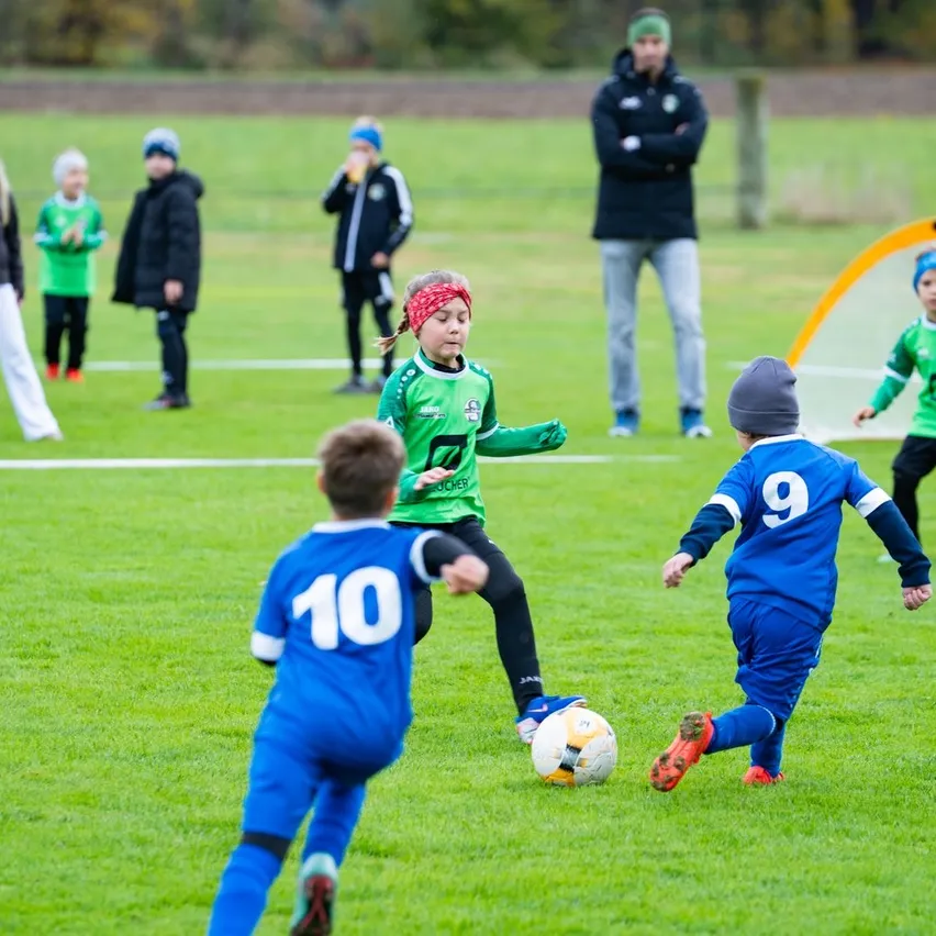 Eine Gruppe von Kindern spielt Fußball auf einem Rasenfeld. Ein Mädchen in einem grünen Shirt läuft auf den Ball zu.