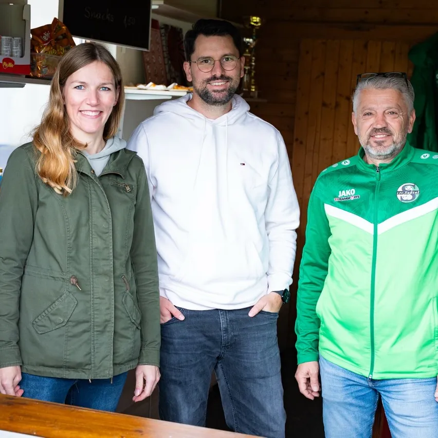 Drei Personen stehen in einer Küche und lächeln für ein Foto. Eine Frau in einer grünen Jacke, ein Mann in einer weißen Kapuzenjacke und ein Mann in einer grünen Jacke mit Logo.