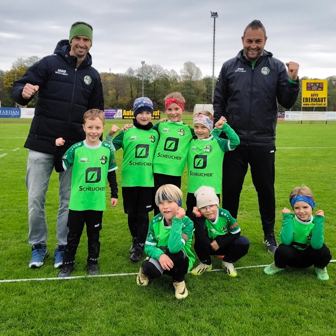Eine Gruppe von Kindern und zwei Männern posiert für ein Foto auf einem Fußballfeld. Die Kinder tragen grüne Uniformen mit dem Wort 'Scheucher' darauf. Die beiden Männer tragen schwarze Jacken und lächeln.